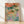 Impressionist-style framed poster depicting a dreamy wildflower garden path with terracotta orange blooms, sage green foliage, and a teal blue stream winding through soft lavender and cream tones.