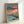 Impressionist-influenced woodblock art of an autumn landscape with a peaceful lake reflecting fall trees in golden and orange hues, surrounded by grey mountains beneath a soft pink sunset sky.