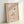 Minimalist framed art print in Japanese woodblock tradition: figure sweeping autumn leaves against cream background with charcoal and tan brushwork creating serene seasonal mood.