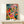 Contemporary botanical interior poster in Bauhaus design featuring flowering plants, leafy arrangements, and a figure in a sanctuary-like setting with golden yellow, emerald, and coral accents.