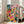 Mid-century modern art print of a flower shop or plant-filled interior with a woman, potted blooms, and decorative shelves in vibrant emerald green, coral pink, and warm cream tones.