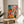 Modern botanical sanctuary art depicting a woman with flowering plants and potted greenery on shelves, rendered in warm coral pink, golden yellow, orange-red, and deep emerald green.