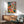 Contemporary botanical wall art in mid-century modern style showing a figure surrounded by blooming flowers and potted plants on shelves in rich coral, golden yellow, and emerald tones.