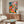 Modern botanical art print featuring a woman amid flowering plants, leafy arrangements, and decorative shelving in bold Bauhaus-influenced colors of coral, emerald, golden yellow, and cream.