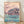 Serene Japanese woodblock-style poster depicting pale pink cherry blossoms along a turquoise river with a traditional teahouse and small boat, rendered in soft cream and sage green tones.