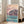 Framed wall art in Japanese woodblock tradition: serene sakura landscape with a boat on turquoise water, teahouse building, and flowering trees in soft pink and cream tones.