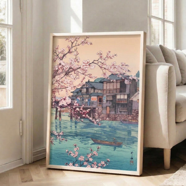 Framed wall art in Japanese woodblock tradition: serene sakura landscape with a boat on turquoise water, teahouse building, and flowering trees in soft pink and cream tones.