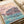 Japanese-style poster art capturing a quiet moment at a sakura-lined river with a small boat near a traditional building, rendered in serene pale pink, cream, and turquoise palette.
