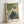 Soft, luminous garden scene with a solitary woman among flowering plants and trees, painted in gentle greens and pale blues with warm ochre accents in an Impressionist aesthetic.