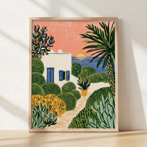 Framed coastal cottage print featuring a Mediterranean-style house with palm trees, cacti, and flowering plants along a path leading to the ocean at sunset, painted in coral pink, deep blue, sage green, and golden yellow tones.