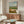 Contemplative watercolor poster depicting a steaming coffee cup, eyeglasses, and newspaper on a windowsill with a leisurely landscape view in rust orange, blue, and cream hues.