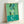Wall-mounted poster displaying colorful geometric cutout shapes in warm hues set against a deep green backdrop, creating a balanced modernist composition.