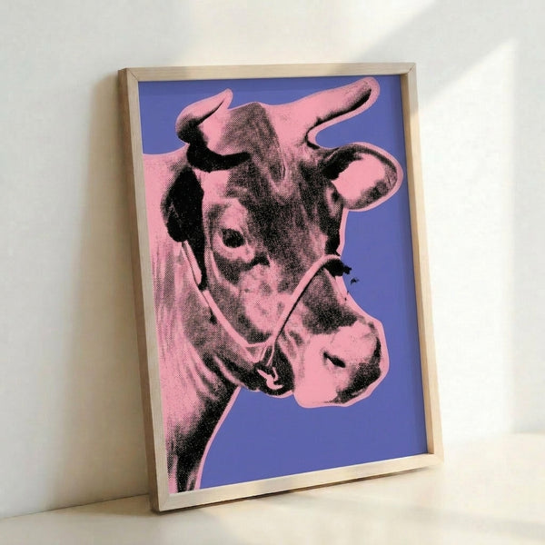 Colorful pop art cow design with pink subject matter contrasting against a rich purple backdrop, shown as a framed poster on a wall.