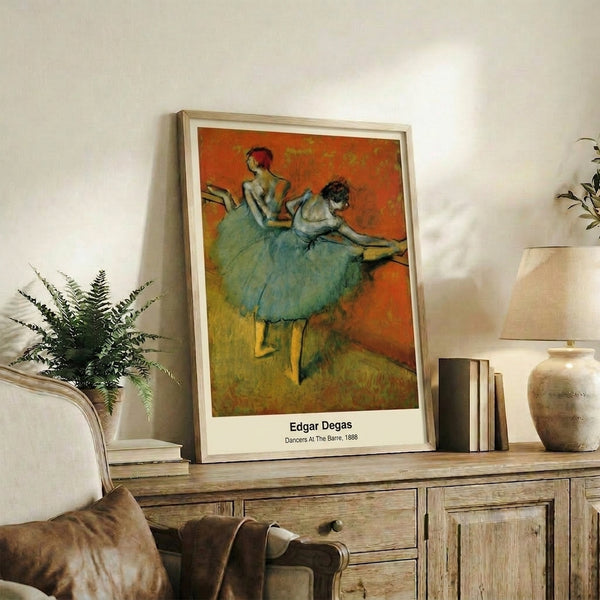Framed Impressionist artwork of dancers stretching at the barre in soft cream and ochre tones, evoking a contemplative moment in the ballet studio.