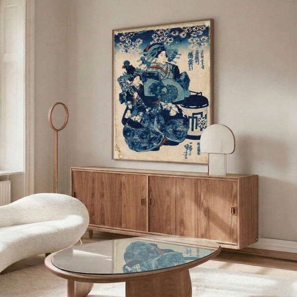 Framed wall art depicting a refined geisha figure in cream, pale blue, and deep indigo kimono with a child companion amid cherry blossom branches, rendered in classic Japanese woodblock style.