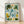 Watercolor poster of an elephant mother and calf surrounded by lush jungle foliage, palm fronds, and blooming flowers in soft sage green, cream, and golden yellow tones, framed and displayed on a wall.