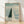Surreal artwork featuring a solitary figure walking on an enormous piano keyboard boardwalk stretching toward a dreamy horizon where clouds meet water, rendered in soft sage green, cream, and pale blue tones.