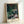 Soft and dreamy Impressionist artwork depicting spring flowers and water lilies beneath a weeping willow, with shimmering reflections in cool greens and whites.