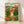 Folk art illustration of a woman in a wildflower meadow gathering blooms into a basket, with mountains in the distance and a warm cottagecore palette of orange, magenta, forest green, and cream.