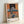 Contemporary art print showing a fragmented seated figure with geometric furniture and glowing light source, executed in a moody palette of slate blue, cream, and burnt orange creating psychological tension.