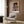 Abstract modern art print featuring a solitary figure with surrounding furniture and suspended light, composed in muted burnt orange, slate blue, and dark brown tones suggesting quiet unease.
