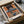 Contemporary poster art with fragmented figure, table, chair, and glowing light bulb in an abstract composition of warm and cool tones—burnt orange, cream, and slate blue—evoking existential introspection.