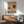Modern minimalist poster depicting a contented frog in a bubble-filled tub with surrounding potted plants and warm terracotta accents, in soothing cream and sage green hues.