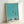 Contemporary minimalist framed poster showing a contemplative moment of a girl waiting at traffic lights on an urban street, with bicycle and lamp post, in muted turquoise and cream.