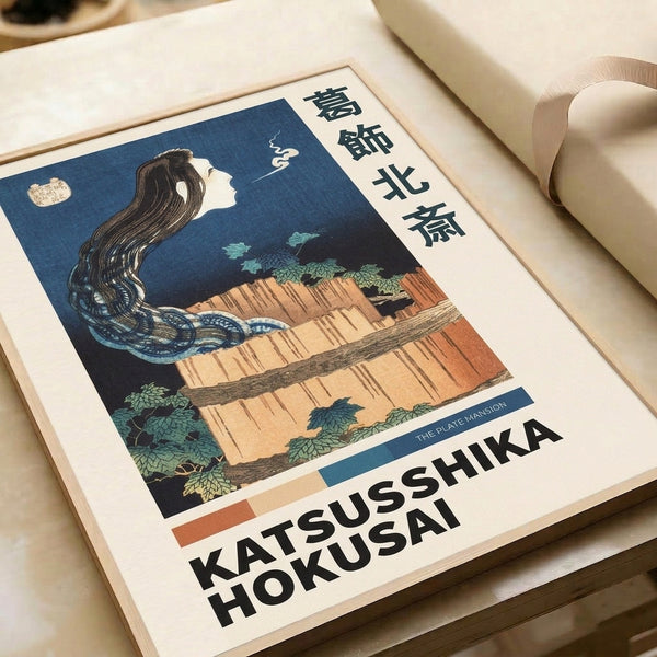 Traditional ukiyo-e framed print with a serene woman positioned before dramatic waterfall scenery and leafy vegetation in moody indigo and cream palette.