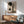 May include: Modern living room with sage green armchair, wooden sideboard, and white table lamp. Large framed abstract art print featuring black circles, geometric rectangles, and silhouetted pine trees in neutral beige and black tones. Natural sunlight creates dramatic shadows across white wall. Mid-century modern furniture with warm wood tones complements the minimalist artwork's organic and geometric elements.