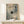 May include: Framed poster with abstract watercolor background in blue, pink and cream tones featuring a vinyl record design with song lyrics arranged in a spiral pattern around the center. Black silhouette of a turntable arm extends from the record. Text placeholders show "Song Name," "Artist Name," and custom text areas. Wood frame sits on light wooden shelf next to small white wrapped gift with natural twine and evergreen sprig against cream wall.