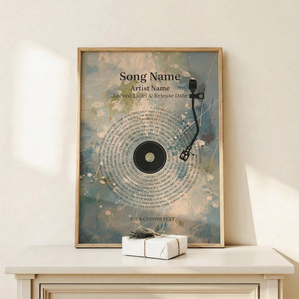 May include: Framed poster with abstract watercolor background in blue, pink and cream tones featuring a vinyl record design with song lyrics arranged in a spiral pattern around the center. Black silhouette of a turntable arm extends from the record. Text placeholders show "Song Name," "Artist Name," and custom text areas. Wood frame sits on light wooden shelf next to small white wrapped gift with natural twine and evergreen sprig against cream wall.