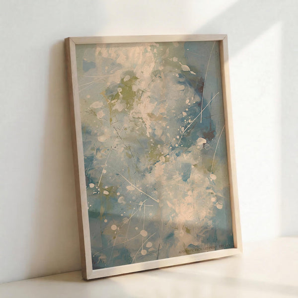 Abstract expressionist artwork in light wooden frame leaning against white wall. Soft blue, green, and cream paint splatters blend with delicate white dots and thin crossing lines throughout. The dreamy, ethereal composition has a watercolor-like quality with organic paint drips and gentle color transitions creating a peaceful, contemporary aesthetic.