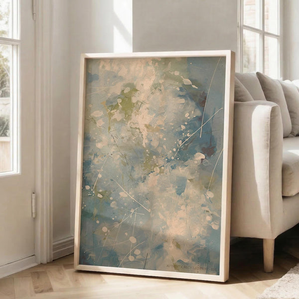 May include: Large framed abstract expressionist artwork leaning against white wall next to beige upholstered chair. The painting features soft blue, green, and peachy pink watercolor-like washes with white circular dots and thin linear elements scattered throughout. Natural wood frame surrounds the piece, which appears to be approximately 2-3 feet tall. Bright natural light streams through nearby windows onto hardwood floors in this minimalist living room setting.