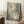 May include: Large framed abstract expressionist artwork leaning against white wall corner, featuring blue and green watercolor washes with white paint splatters and linear scratches throughout. Natural wood frame, positioned on white textured rug beside wooden side table with white coffee cup and saucer. Soft natural lighting creates geometric window shadows on wall.
