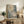 May include: Abstract expressionist artwork in light wood frame displaying blue, green, and peachy pink paint splatters with white linear elements, positioned on rustic wooden sideboard alongside fern plant, ceramic vessels, and books in neutral-toned living room setting with cream upholstered chair visible.
