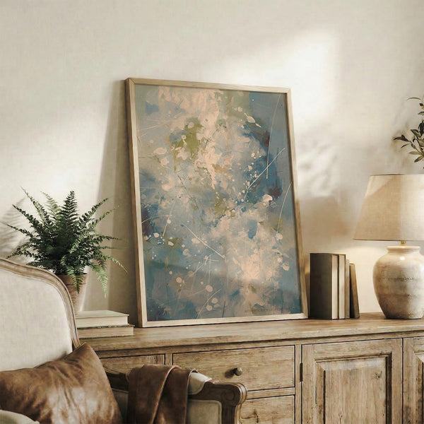 May include: Abstract expressionist artwork in light wood frame displaying blue, green, and peachy pink paint splatters with white linear elements, positioned on rustic wooden sideboard alongside fern plant, ceramic vessels, and books in neutral-toned living room setting with cream upholstered chair visible.