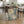 May include: Abstract expressionist artwork with blue, green, and cream splashes and splatters, displayed in light wood frame leaning against wooden dining table with hairpin legs. Kitchen setting features wicker chairs, spider plant, wine bottle, and white cabinets. Artwork shows organic flowing paint textures with scattered dots and gestural brushwork on light background.
