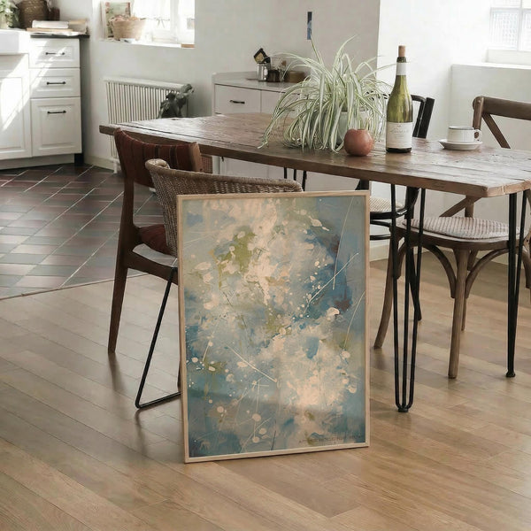 May include: Abstract expressionist artwork with blue, green, and cream splashes and splatters, displayed in light wood frame leaning against wooden dining table with hairpin legs. Kitchen setting features wicker chairs, spider plant, wine bottle, and white cabinets. Artwork shows organic flowing paint textures with scattered dots and gestural brushwork on light background.