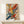 May include: Abstract expressionist artwork with vibrant paint splatters and brushstrokes in yellow, blue, red, orange, and green on white background, displayed in light wood frame on white shelf next to small wrapped gift with evergreen sprig against cream wall