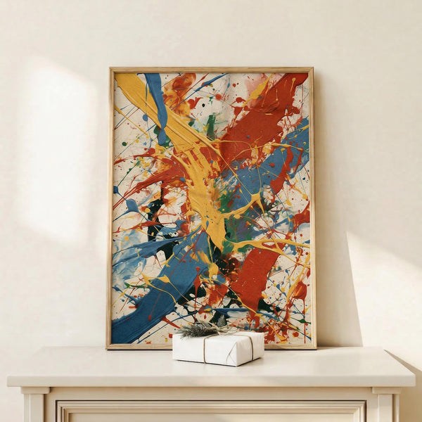 May include: Abstract expressionist artwork with vibrant paint splatters and brushstrokes in yellow, blue, red, orange, and green on white background, displayed in light wood frame on white shelf next to small wrapped gift with evergreen sprig against cream wall
