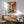 May include: Colorful abstract expressionist artwork with vibrant yellow, red, and blue paint splatters and brushstrokes in wooden frame, hanging above mid-century modern walnut credenza with white ceramic table lamp, next to sage green upholstered armchair with wooden arms in bright sunny room with white walls and natural lighting creating soft shadows.