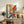 May include: Large framed abstract expressionist artwork with vibrant paint splatters and brushstrokes in yellow, blue, red, and orange on white canvas, displayed on rustic wooden sideboard alongside fern plant, books, and ceramic vases in neutral-toned living room setting with beige chair and pillows.