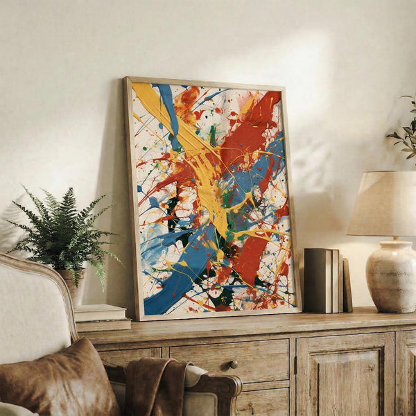 May include: Large framed abstract expressionist artwork with vibrant paint splatters and brushstrokes in yellow, blue, red, and orange on white canvas, displayed on rustic wooden sideboard alongside fern plant, books, and ceramic vases in neutral-toned living room setting with beige chair and pillows.
