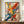 May include: Large framed abstract expressionist artwork with vibrant paint splatters and drips in yellow, red, blue, green, and black against white background, displayed on wooden sideboard next to white ceramic vase and stack of books in elegant room with cream walls and crown molding.