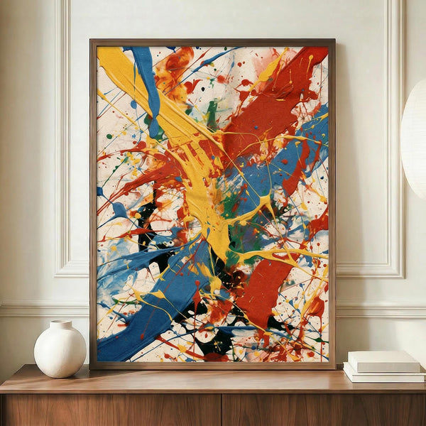 May include: Large framed abstract expressionist artwork with vibrant paint splatters and drips in yellow, red, blue, green, and black against white background, displayed on wooden sideboard next to white ceramic vase and stack of books in elegant room with cream walls and crown molding.