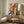 May include: Vibrant abstract expressionist artwork with bold red, yellow, and blue paint splatters and brushstrokes on white canvas, displayed in thin white frame on wooden sideboard next to cream ceramic table lamp. Modern minimalist room setting with beige textured chair, round wooden side table, and neutral walls.