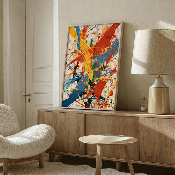 May include: Vibrant abstract expressionist artwork with bold red, yellow, and blue paint splatters and brushstrokes on white canvas, displayed in thin white frame on wooden sideboard next to cream ceramic table lamp. Modern minimalist room setting with beige textured chair, round wooden side table, and neutral walls.