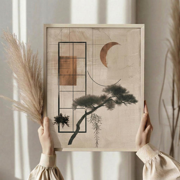 May include: Person in beige clothing holding framed abstract art print featuring geometric rectangular grid overlaying stylized pine tree branch with green foliage, bronze crescent moon, and textured beige background. One hand also holds dried pampas grass stems. Natural lighting with white curtained window backdrop creates soft, minimalist styling scene.