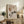May include: Framed abstract artwork displaying geometric rectangles, crescent moon, and stylized pine tree in earth tones on neutral background, positioned on rustic wooden sideboard alongside fern plant, books, ceramic vase, and throw pillows in cozy living room setting with warm natural lighting.